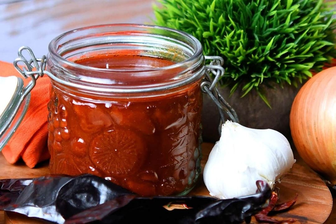 A glass jar of red sauce sits on a wooden surface next to a head of garlic, a whole onion, dried chilies, a green plant, and an orange cloth.