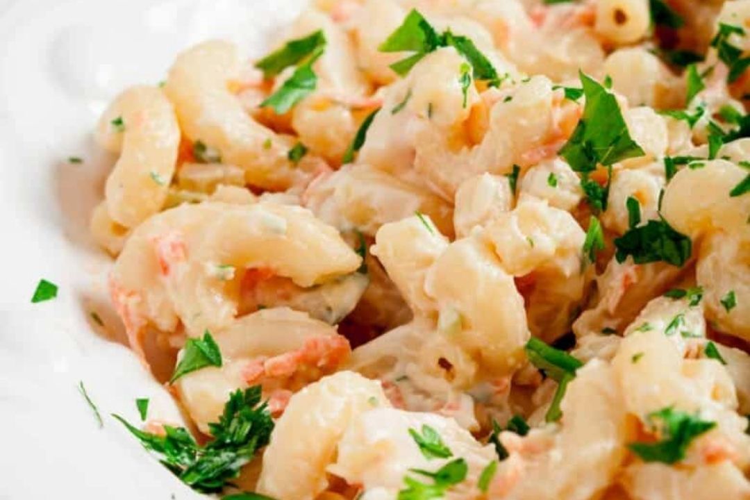 Close-up of creamy macaroni salad with visible pieces of pasta, chopped parsley, and small bits of vegetables on a white plate—an ideal Make-Ahead Labor Day Side for stress-free hosting.