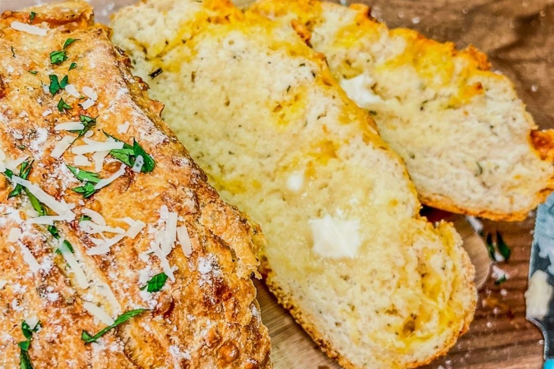 Loaf of bread topped with herbs and cheese, partially sliced to show the soft, buttery interior, on a wooden surface.