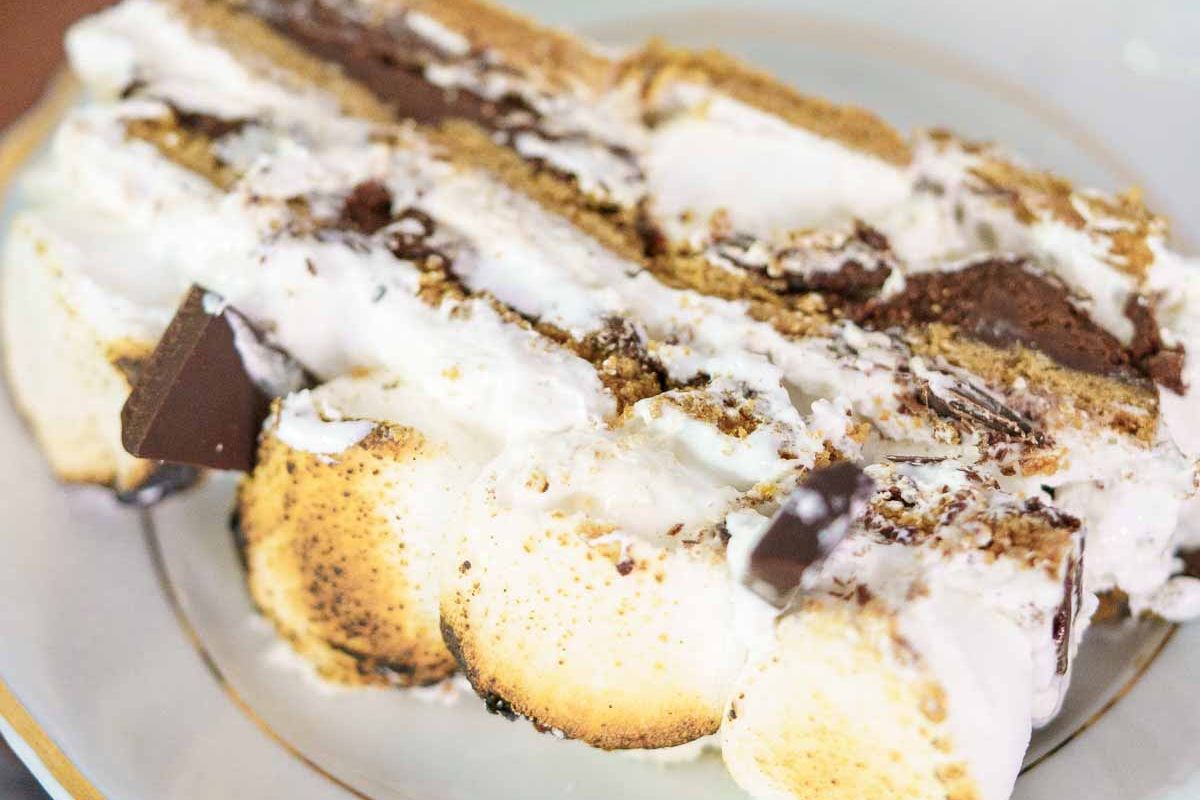A close-up of a slice of layered s’mores cake with toasted marshmallow, chocolate pieces, and cream on a white plate—a delightful treat that’s perfect for summer desserts and easily enjoyed as a gluten free option.