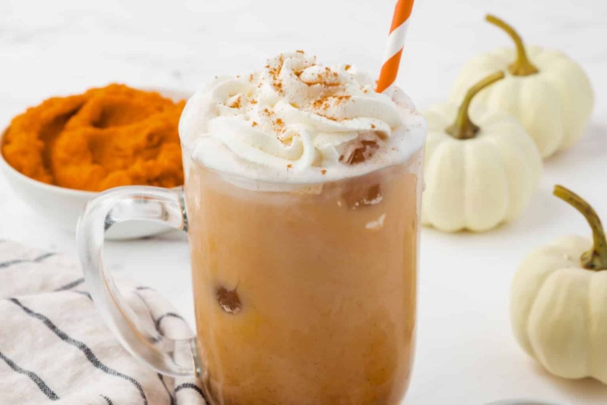 Glass mug of iced coffee with whipped cream and an orange straw, next to a bowl of pumpkin puree, two small white pumpkins, and a striped cloth on a white surface—perfect inspiration for cozy fall recipes during pumpkin season.