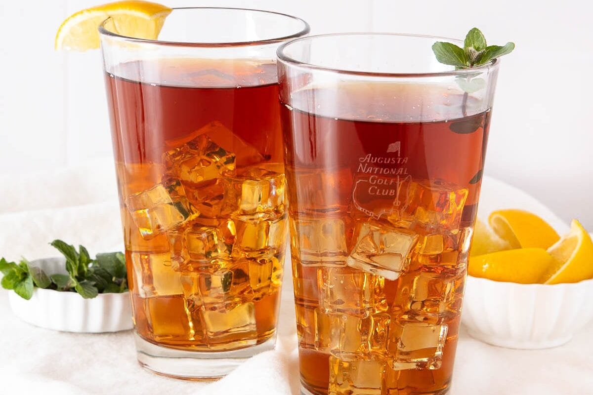 Two glasses of iced tea with lemon wedges and mint garnish, set on a white surface next to a small bowl of lemon slices and a dish of fresh mint—perfect beverages to help you cool off and enjoy a relaxing Labor Day.
