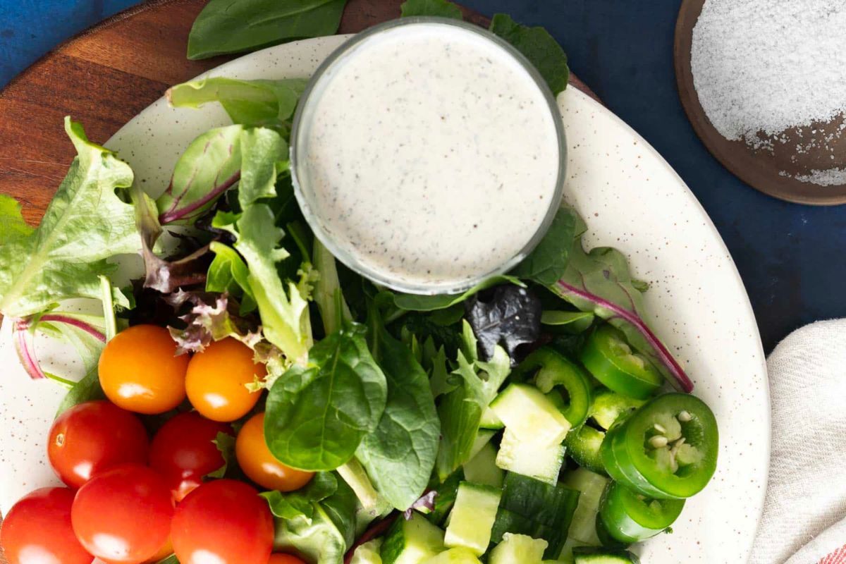 Plate with mixed greens, cherry tomatoes, cucumber, sliced jalapeños, and a small bowl of creamy homemade dressing on a speckled white plate. Salt in a small dish is visible in the corner.