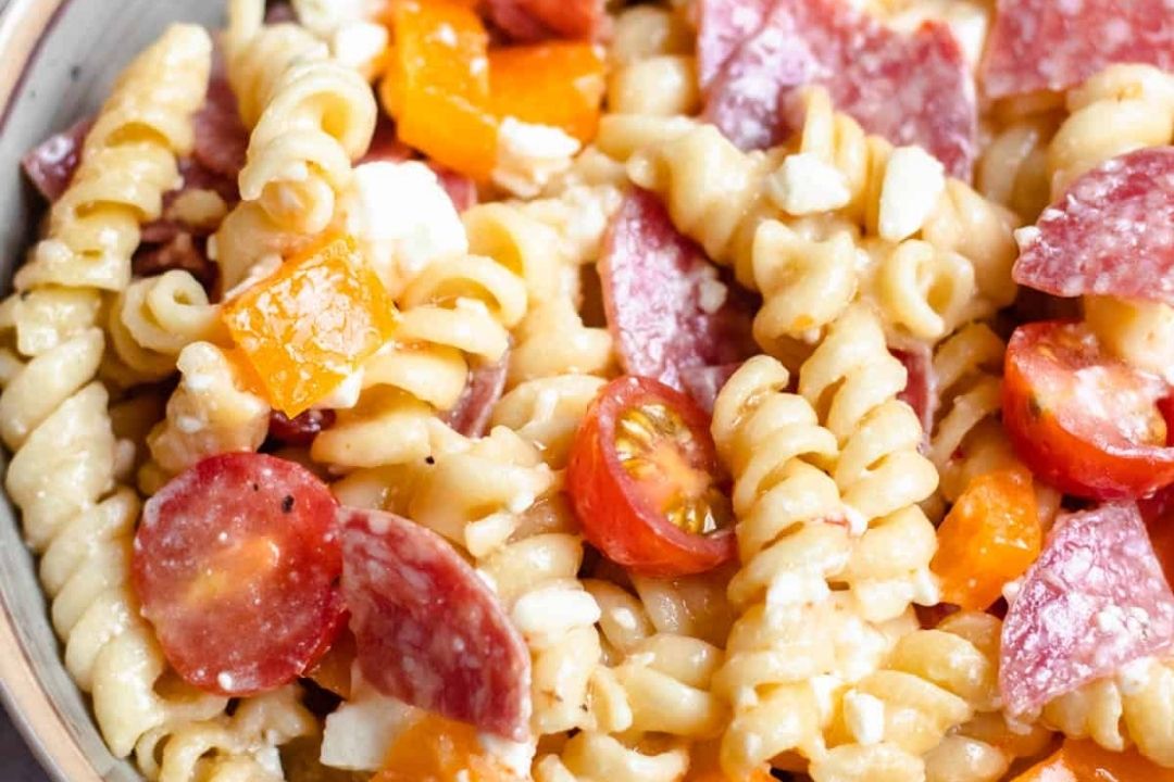 Close-up of rotini pasta salad with cherry tomatoes, diced yellow bell peppers, sliced salami, and crumbled cheese.