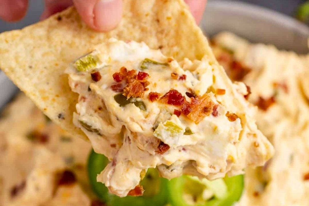 A tortilla chip topped with creamy dip containing chopped green peppers and bacon bits, held above a bowl of the same cold dip—perfect for game day snacking.
