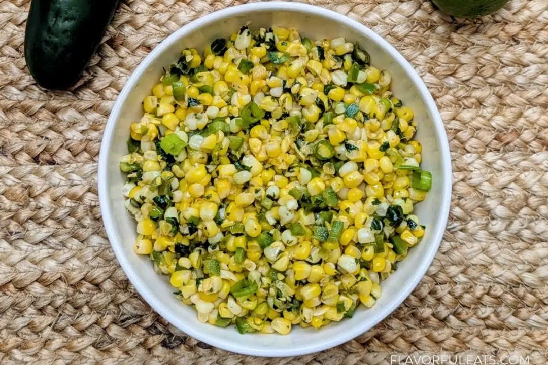 A white bowl filled with a mixture of corn, chopped green onions, and herbs sits on a woven surface.