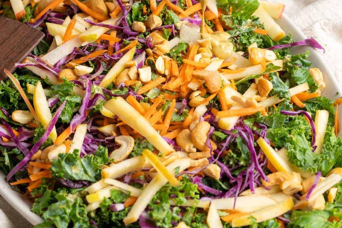 Salad with kale, shredded carrots, purple cabbage, apple slices, and mixed nuts on a white plate—a refreshing option during apple season and a delightful addition to your collection of apple recipes.