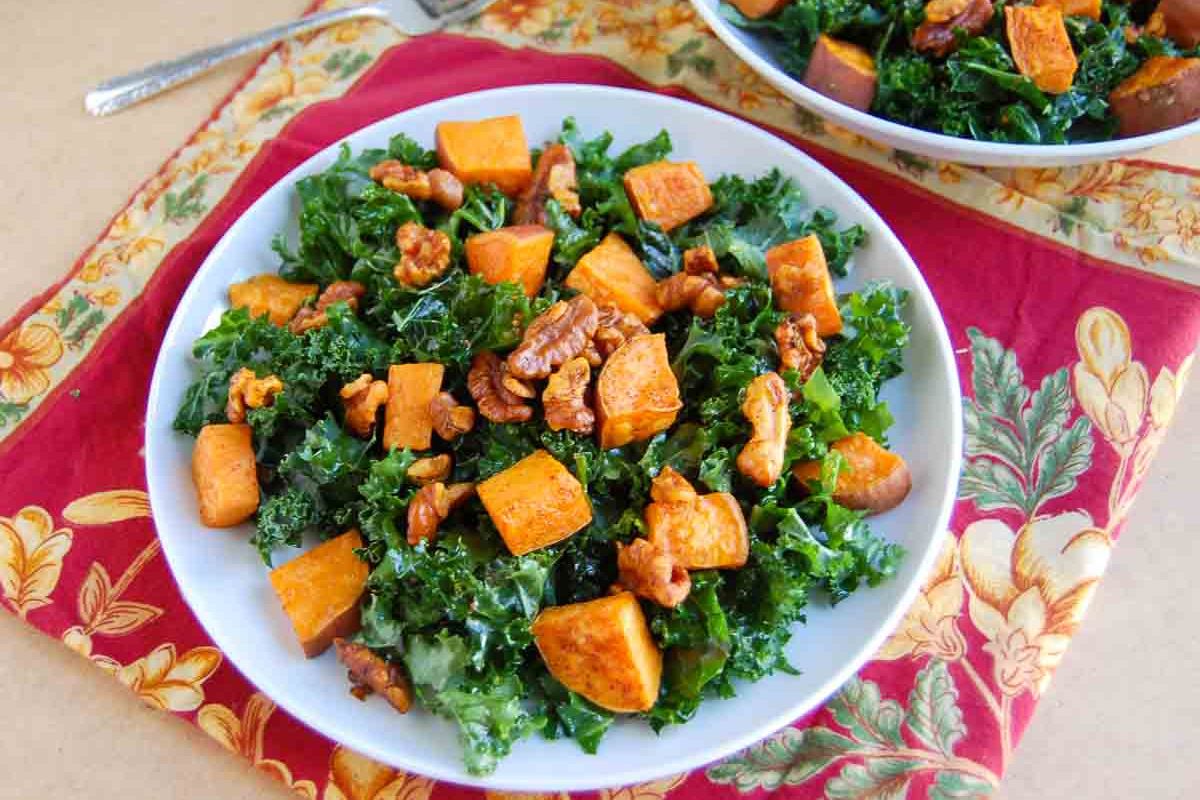 A white bowl filled with kale, roasted sweet potato cubes, and walnuts, placed on a red and yellow floral napkin—one of those travel friendly salads perfect for potlucks or gatherings, as it’s packed with flavor and holds up beautifully on the go.