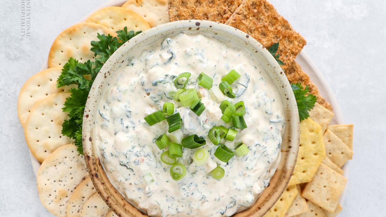 A bowl of creamy cold dip topped with chopped green onions is surrounded by assorted crackers and leafy parsley on a white plate—perfect for game day snacks or party appetizers.