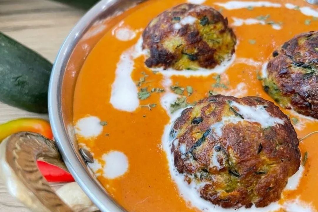Three vegetable kofta balls in a creamy orange curry sauce, garnished with a drizzle of cream and herbs, served in a metal bowl.