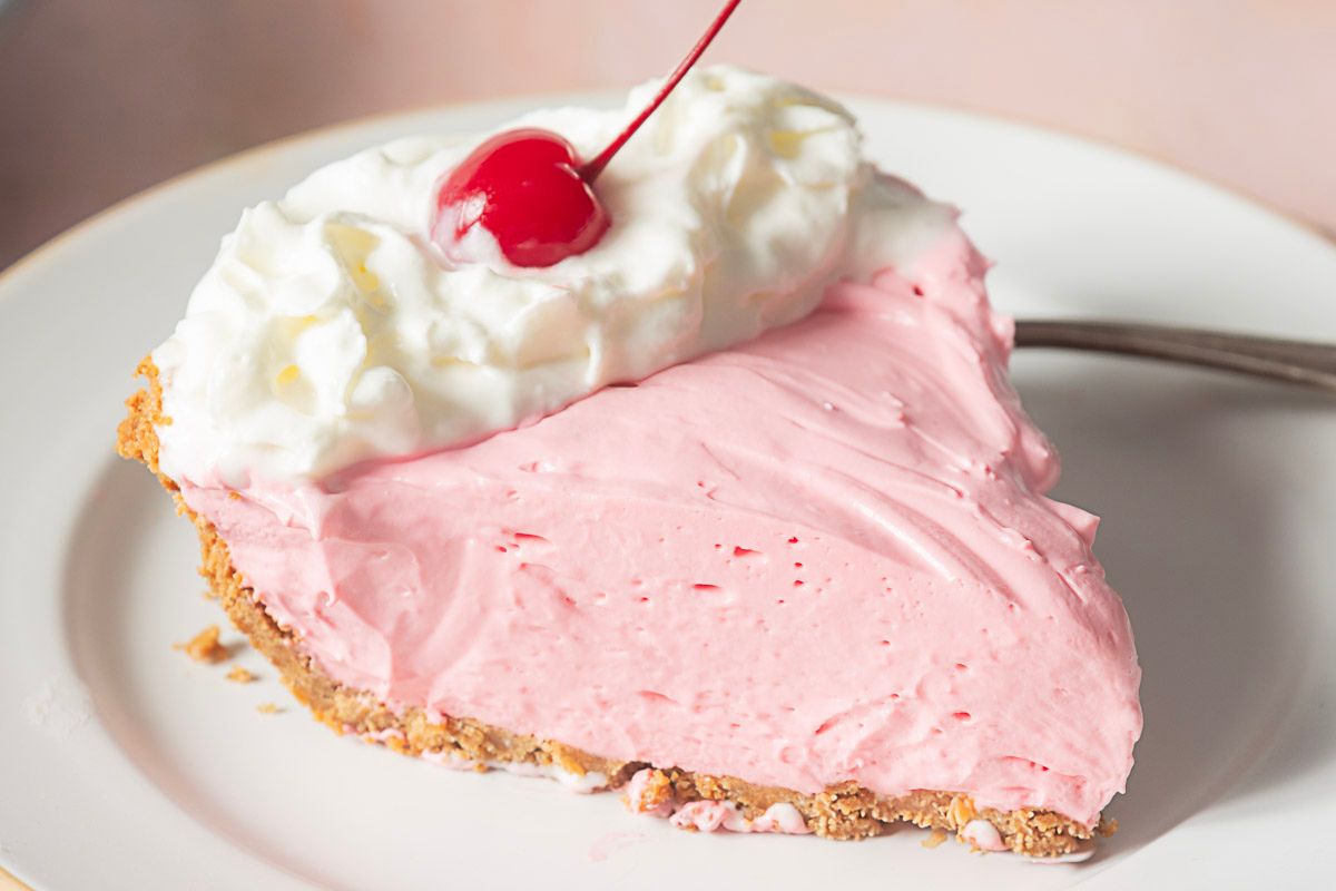 A slice of pink cream pie with a graham cracker crust, topped with whipped cream and a maraschino cherry, on a white plate—the perfect treat for fans of summer desserts and gluten free desserts alike.
