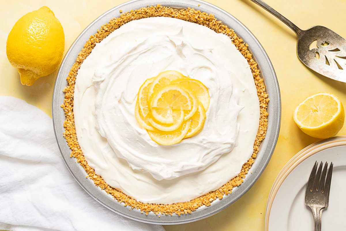 A lemon cream pie with a graham cracker crust is topped with whipped cream and lemon slices, surrounded by fresh lemons, a serving utensil, and plates with a fork on a yellow surface—perfect for those seeking refreshing summer desserts or looking for delicious gluten free options.
