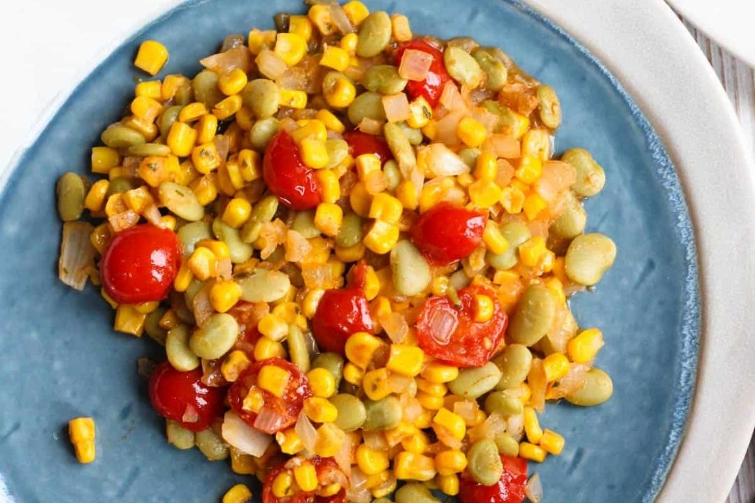A serving of succotash with corn, lima beans, cherry tomatoes, and diced onions on a blue plate.