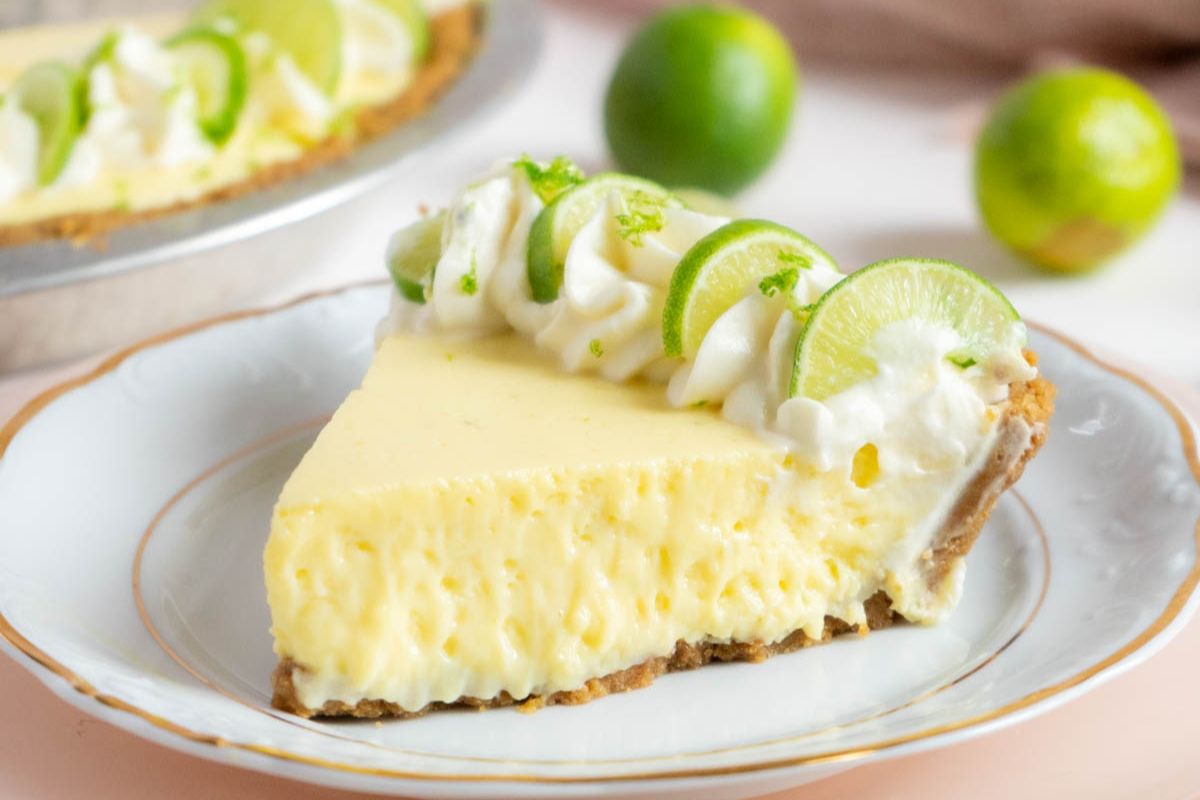 A slice of key lime pie with whipped cream and lime slices on top sits on a white plate; whole limes are in the background—perfect as a refreshing finale for your summer recipes or Labor Day cookout recipes.