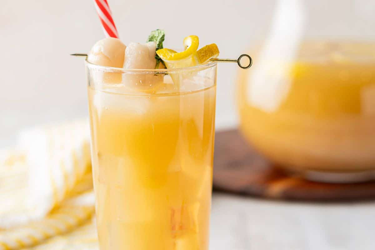 A tall glass of iced yellow drink garnished with lychee, lemon twist, and mint, with a red-striped straw, sits in front of a pitcher of the same drink—a refreshing option perfect for fresh luncheon recipes or guest lunch ideas.