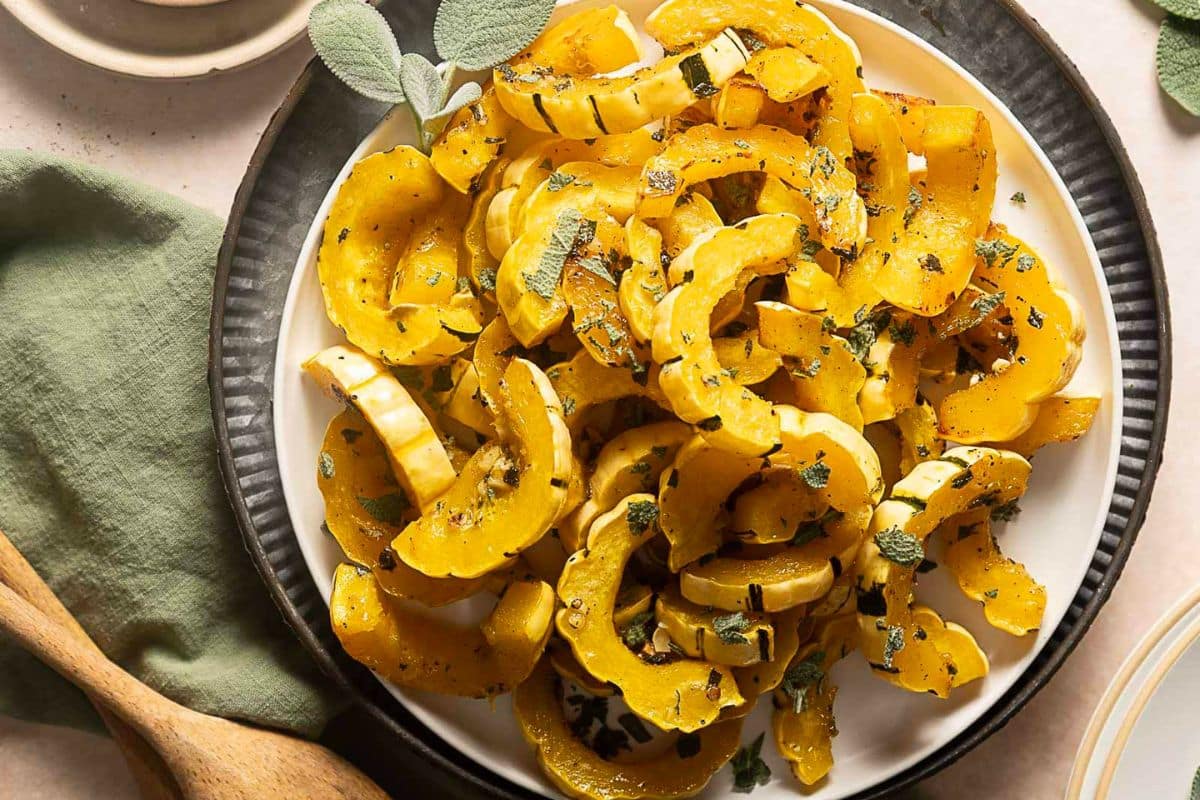 Sliced roasted delicata squash with herbs is arranged on a white plate with a black rim, next to a green napkin and wooden utensils—an inviting example of easy vegetable dishes perfect for creative vegetable meals.
