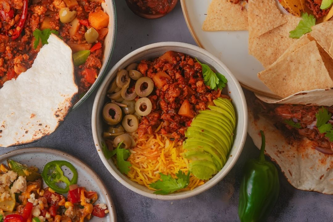 A bowl of yellow rice topped with ground meat, sliced olives, avocado, and cilantro sits near tortilla chips, jalapeño, and various sides.