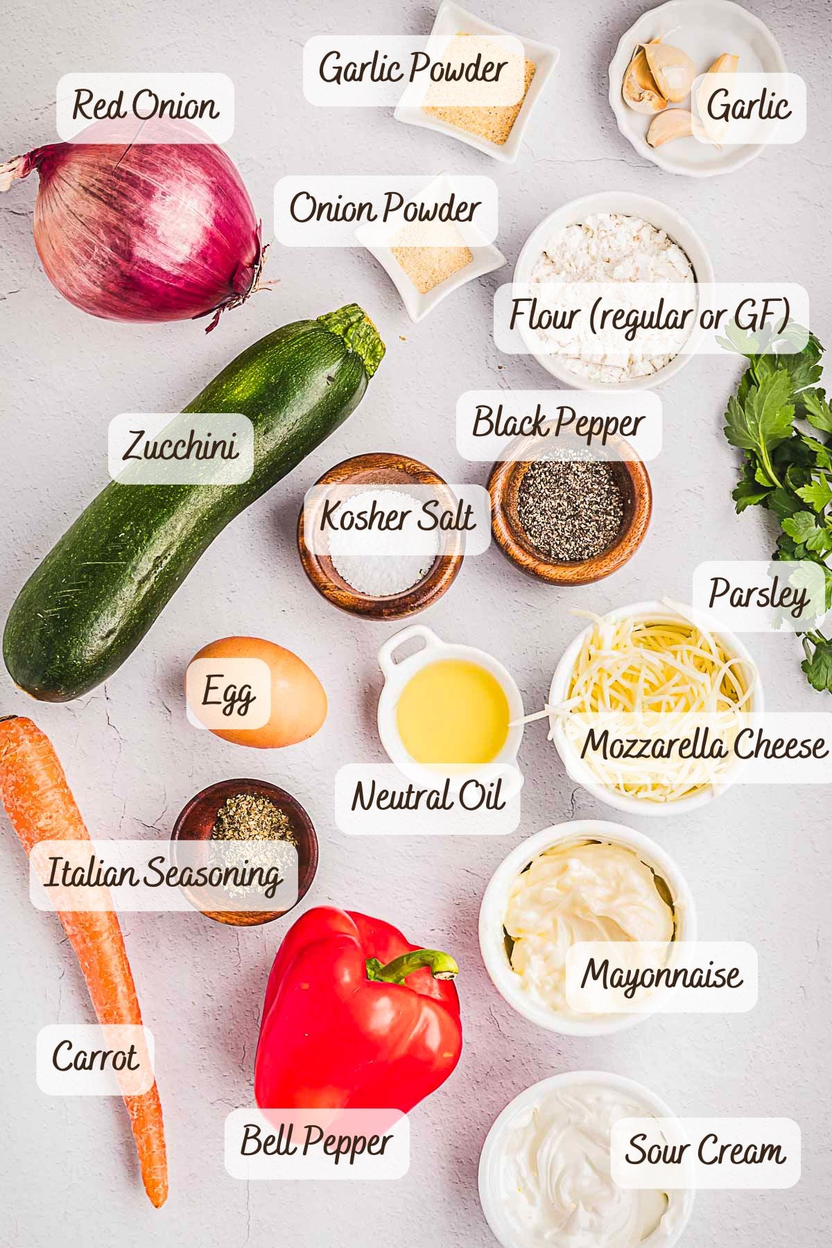 Various labeled ingredients including red onion, garlic, zucchini, carrot, bell pepper, egg, flour, mozzarella cheese, spices, mayonnaise, and sour cream arranged on a white surface—perfect for preparing crispy vegetable fritters with a flavorful garlic sauce.