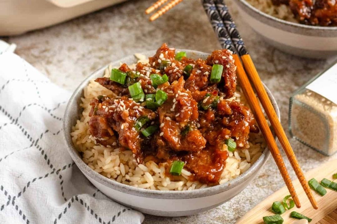 A bowl of rice topped with saucy beef, chopped green onions, and sesame seeds, with chopsticks placed on the bowl’s edge—perfect for cozy fall recipes or as an easy addition to your favorite Crockpot recipes.