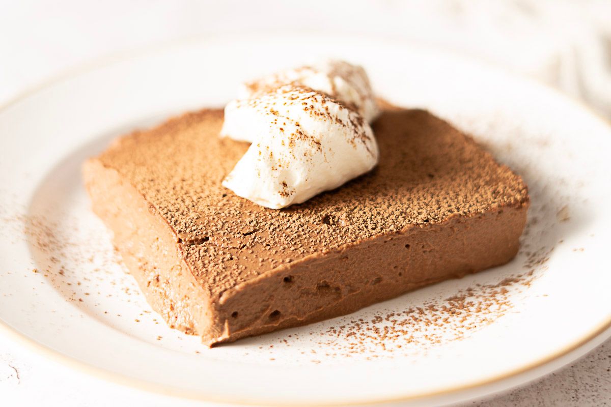A square piece of chocolate mousse topped with whipped cream and dusted with cocoa powder, served on a white plate—a decadent treat perfect for chocolate lovers.