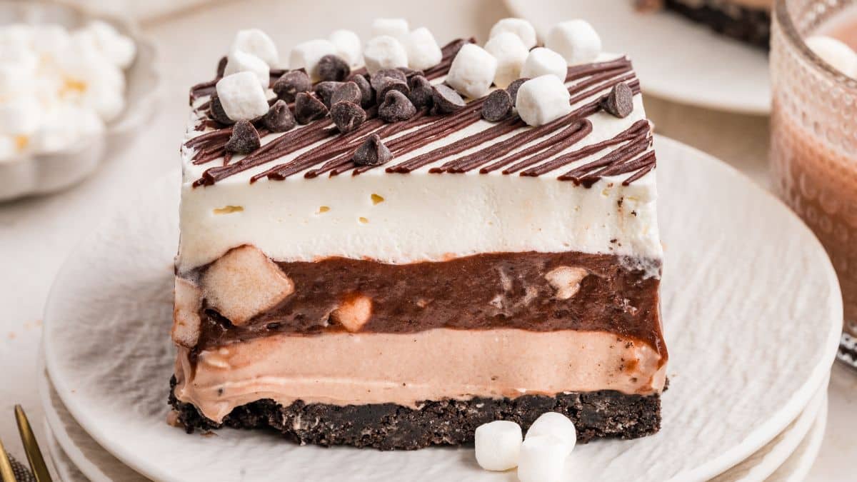 A layered dessert bar with a chocolate cookie crust, light brown and white creamy layers, topped with mini marshmallows, chocolate chips, and a chocolate drizzle—served on a white plate—perfect for chocolate lovers seeking delicious new chocolate desserts.