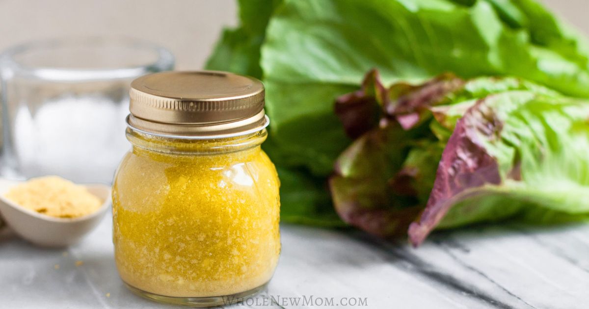 A small glass jar filled with yellow powder sits on a marble surface next to lettuce leaves and a small white spoon with more powder, perfect for creating superior dressings or enhancing your favorite homemade dressings.