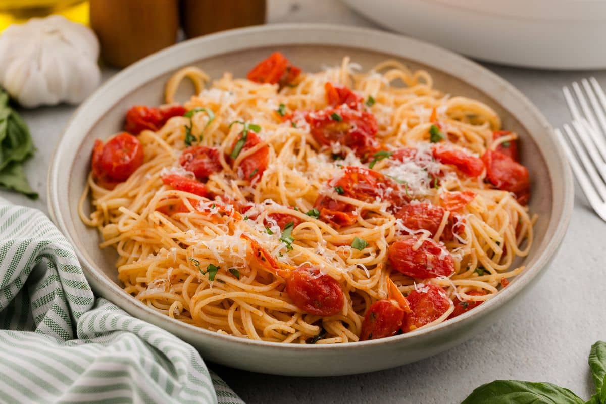 A bowl of spaghetti topped with tomato sauce, cherry tomatoes, grated cheese, and chopped herbs sits on a table with a green-striped napkin—an inviting addition to your collection of fresh and vibrant summer recipes.
