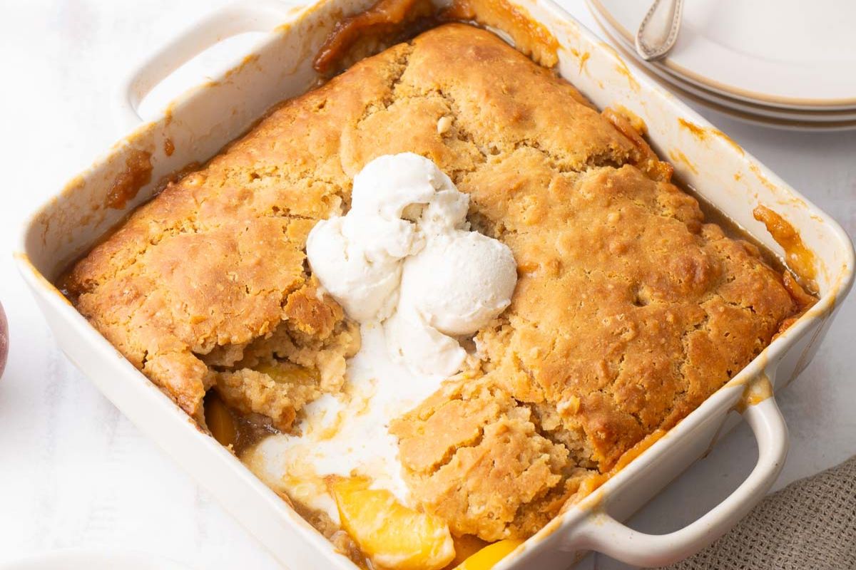 A square dish of peach cobbler with a golden crust, partially scooped to reveal peaches inside, topped with three scoops of vanilla ice cream—this classic treat is perfect for summer desserts and can easily be made gluten free for everyone to enjoy.