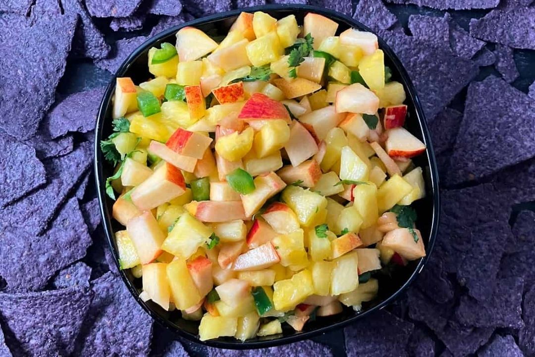 A black bowl filled with diced pineapple, apple, jalapeño, and herbs sits on blue corn tortilla chips.