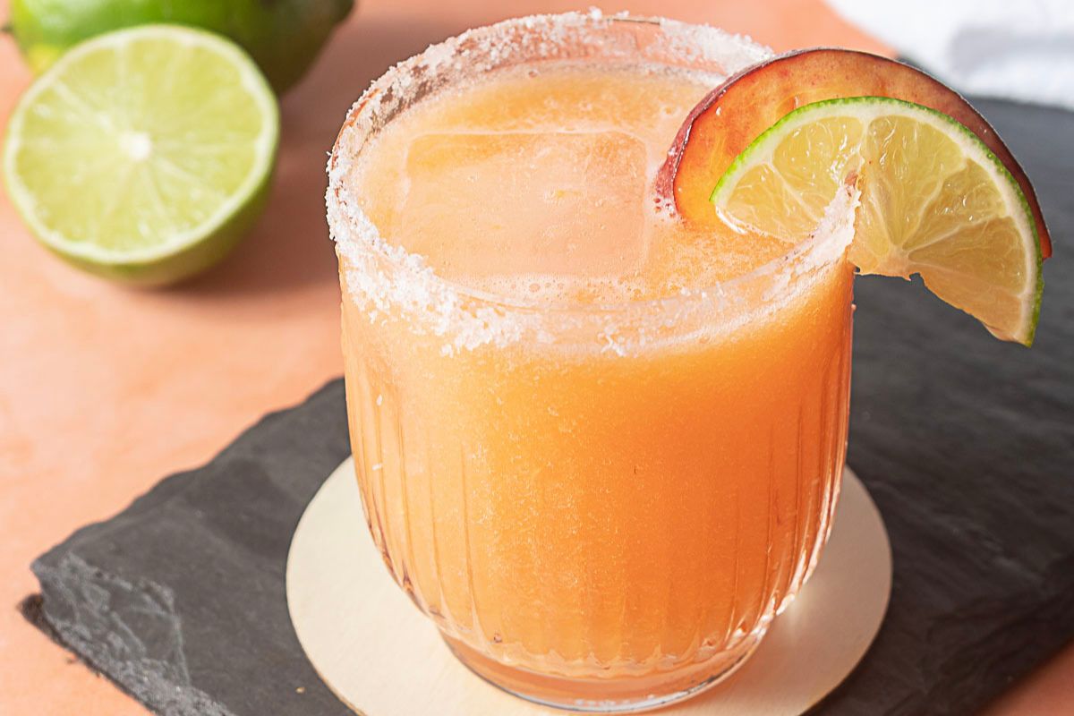 A salt-rimmed glass filled with an orange drink, ice, and garnished with a slice of lime and peach; a halved lime sits in the background—one of the perfect summer refreshments to enjoy on a warm afternoon.