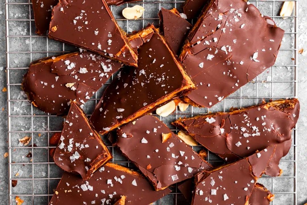 Sliced chocolate-covered toffee pieces topped with flaky salt rest on a wire cooling rack, with some peanuts visible among the pieces—a tempting treat for anyone who loves desserts or is searching for new peanut butter recipes.