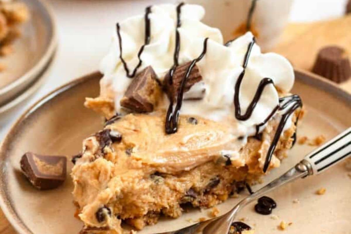 A slice of peanut butter pie, a classic among peanut butter desserts, is topped with whipped cream, chocolate drizzle, and peanut butter cup pieces on a beige plate with a fork.