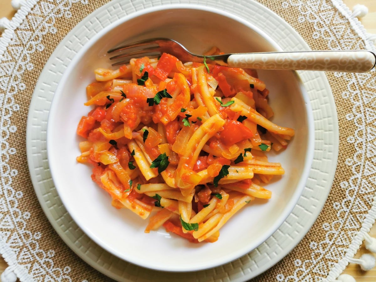 A bowl of pasta with tomato sauce, garnished with chopped herbs, sits on a white plate with a fork on the side, atop a lace-patterned placemat—an ideal addition to any collection of vegetable recipes.