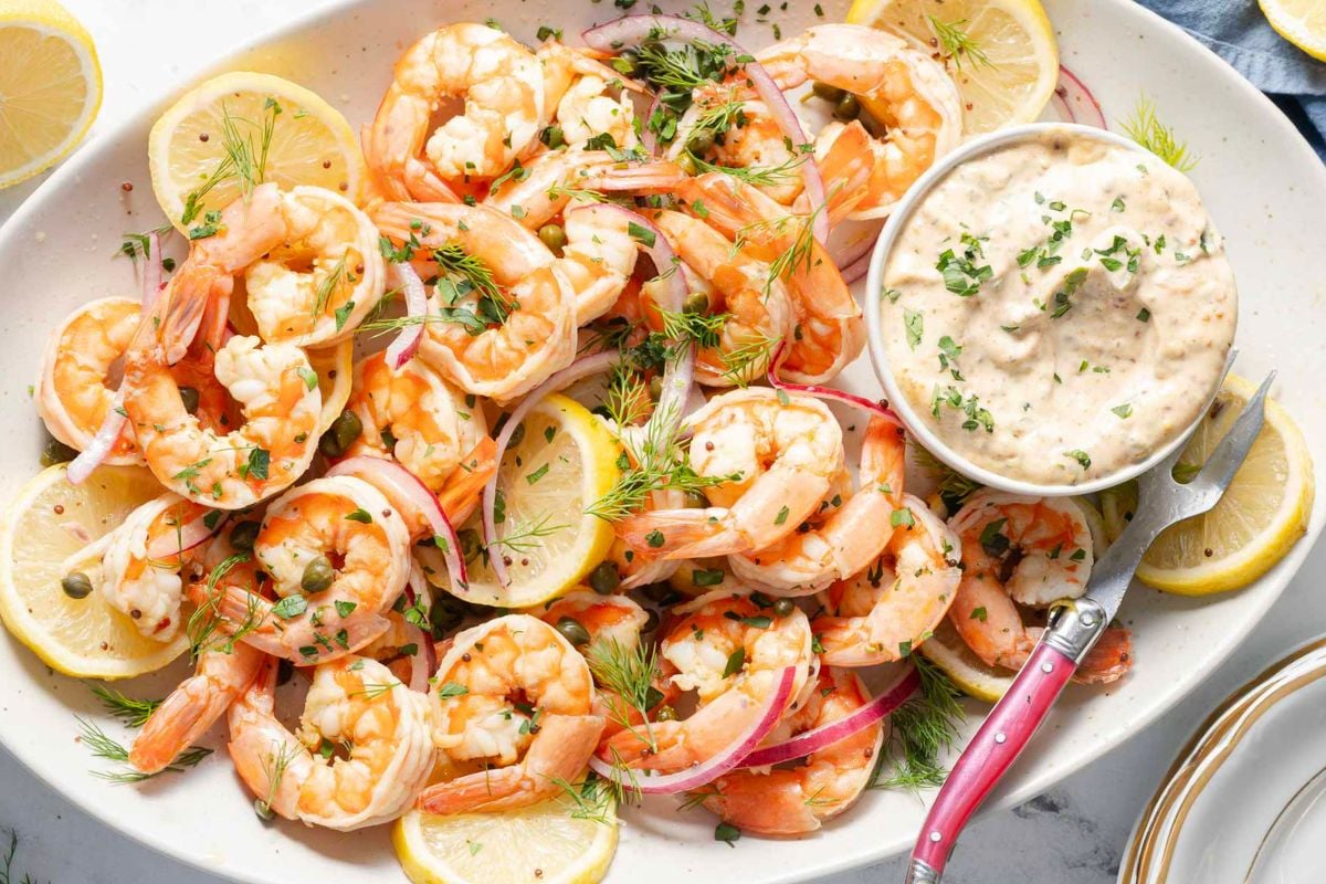Platter of cooked shrimp garnished with dill, red onion, and lemon slices, served with a bowl of dipping sauce and a small fork—an elegant choice for those seeking fresh luncheon recipes or inspiring guest luncheon ideas.