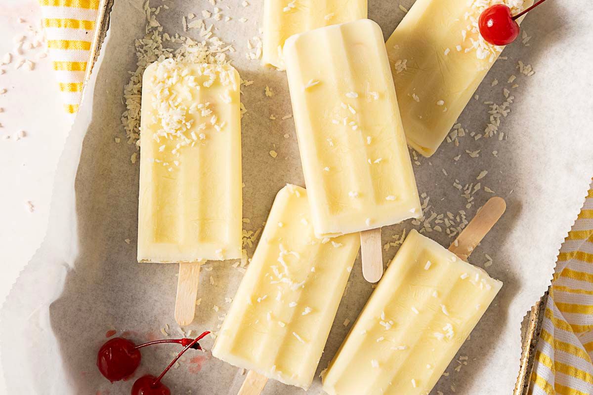 Five creamy white popsicles with shredded coconut on top are arranged on parchment paper with two maraschino cherries nearby—perfect gluten free desserts to enjoy as refreshing summer treats.