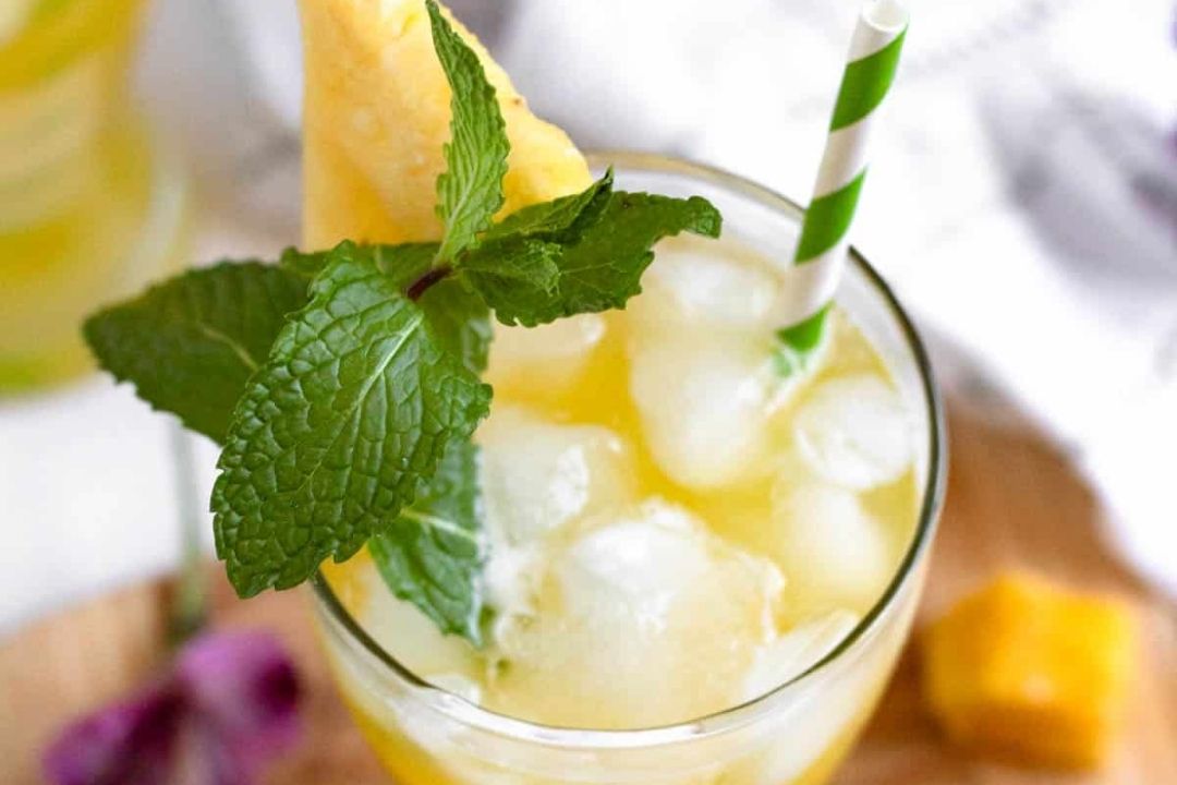 A glass of yellow iced drink garnished with mint leaves, a pineapple wedge, and a green striped straw—perfect to cool off with on Labor Day or anytime you crave refreshing beverages.