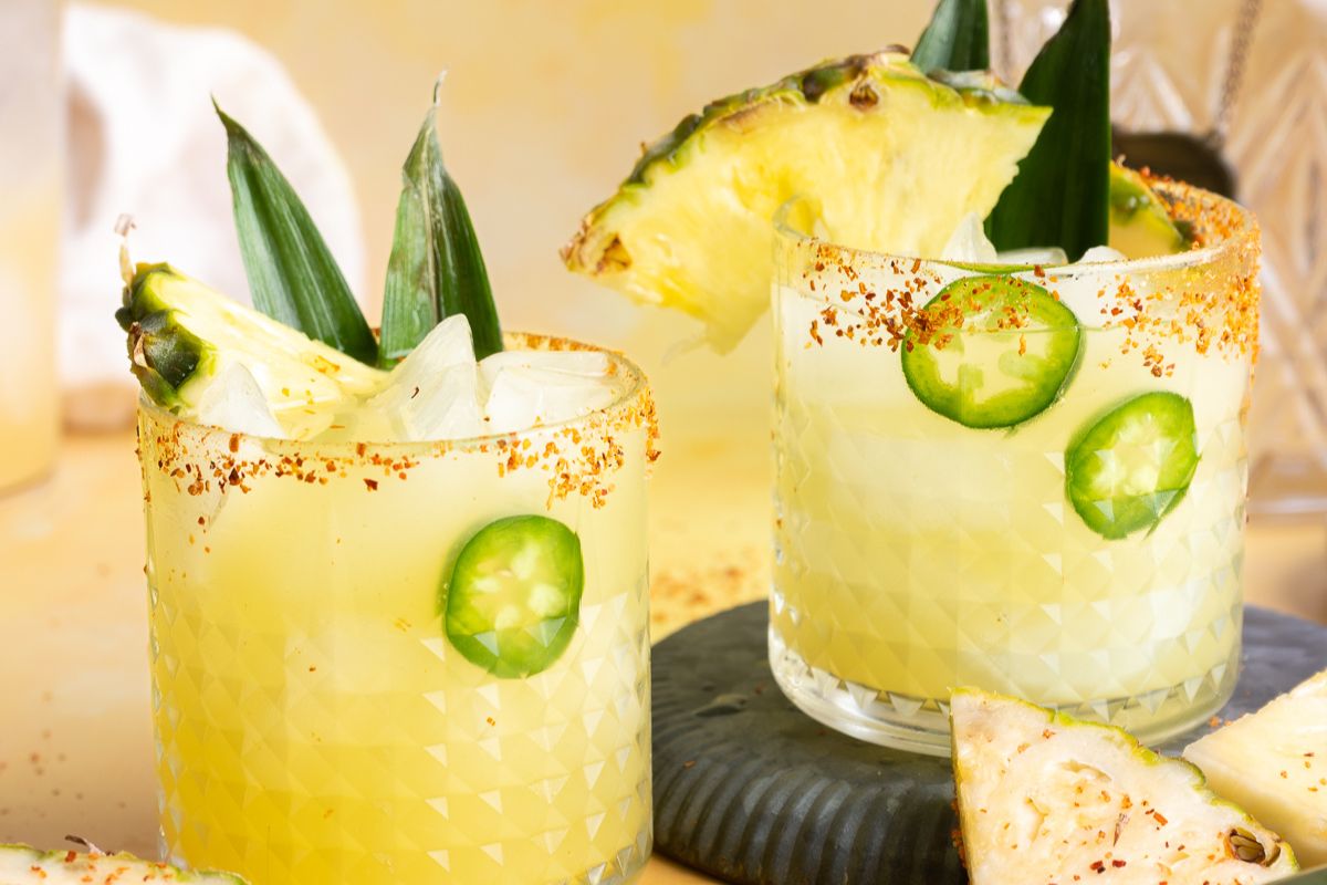 Two glasses of yellow beverages garnished with pineapple wedges, jalapeño slices, ice, and chili powder on the rim, set on a light surface with pineapple pieces nearby—perfect to help you cool off this Labor Day.