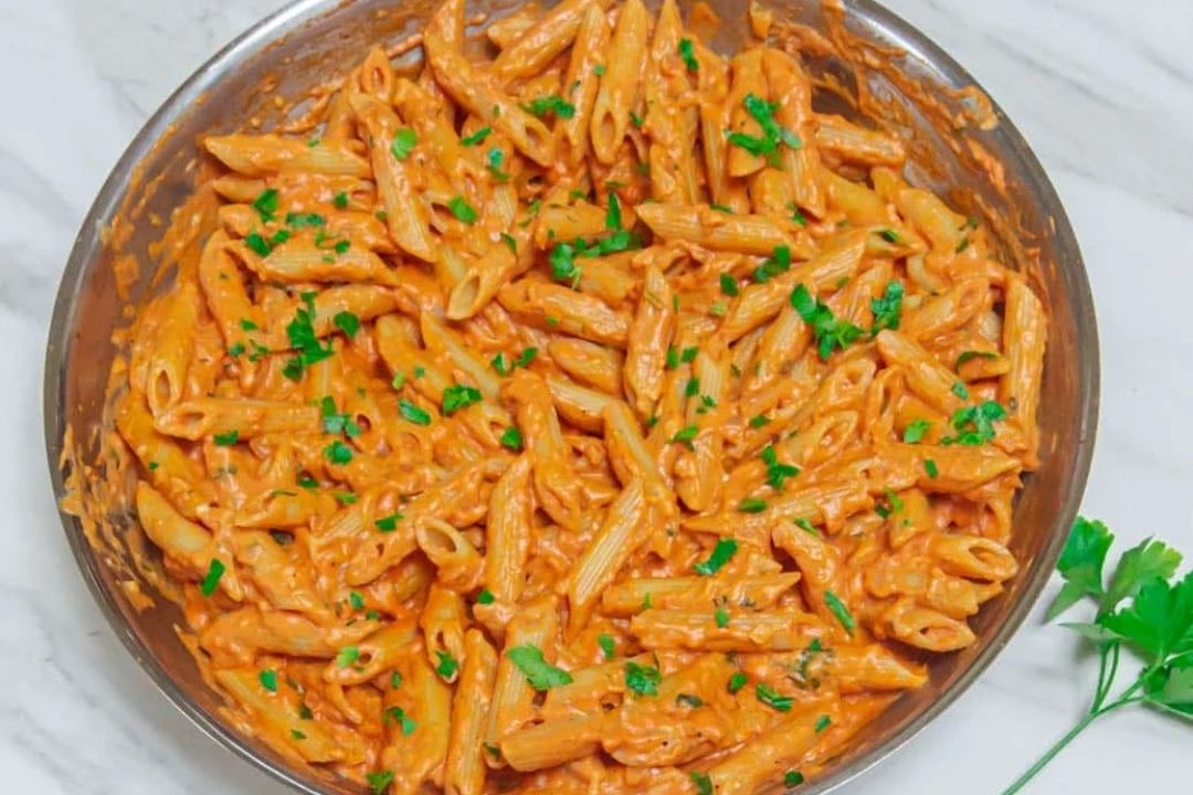 Penne pasta in a creamy tomato sauce, garnished with chopped parsley, served in a round pan on a marble surface.