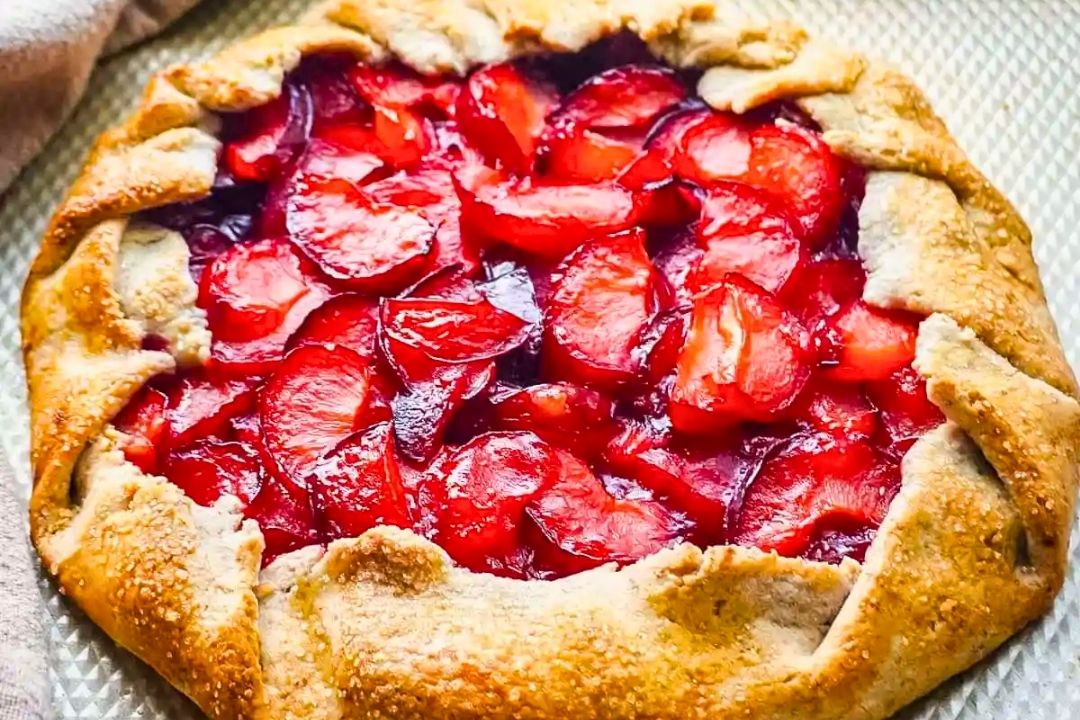 A rustic plum galette with a golden-brown crust and sliced plums arranged in the center, sitting on a textured surface—this centerpiece perfectly captures the essence of summer recipes.