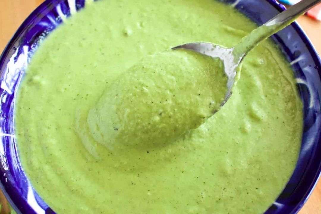 A spoon holds thick, green sauce above a blue bowl filled with the same creamy green mixture.