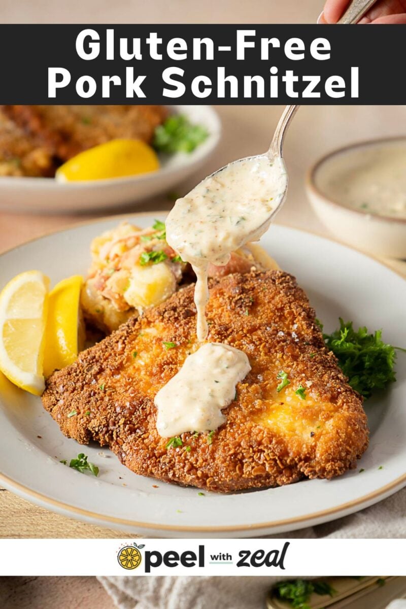 A plate with gluten-free pork schnitzel, creamy sauce being spooned on top, lemon wedges, potato salad, and parsley garnish.