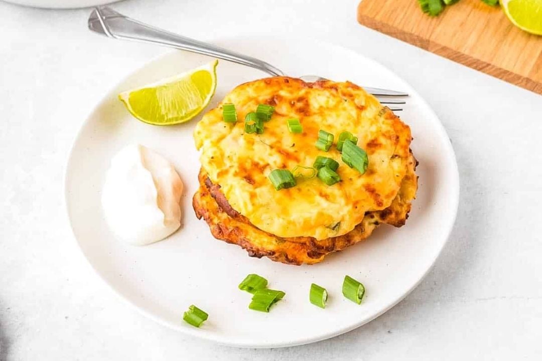 Two cheesy fritters topped with chopped green onions, served on a white plate with a dollop of sour cream, a lime wedge, and a fork.