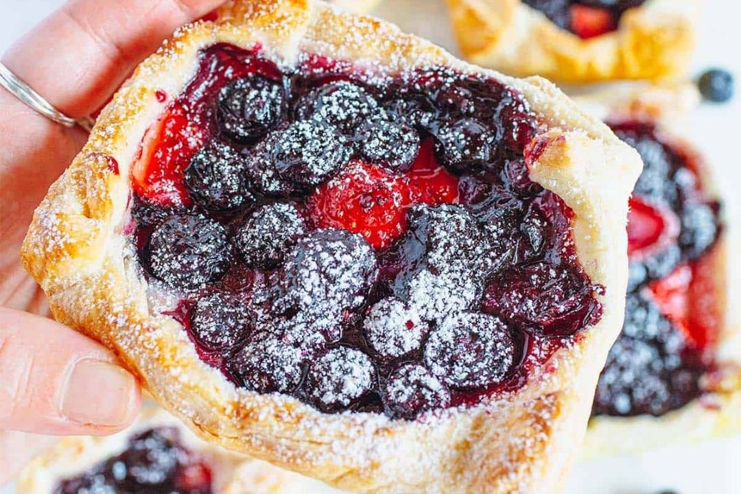 A hand holds a square pastry topped with blueberries, raspberries, and powdered sugar.