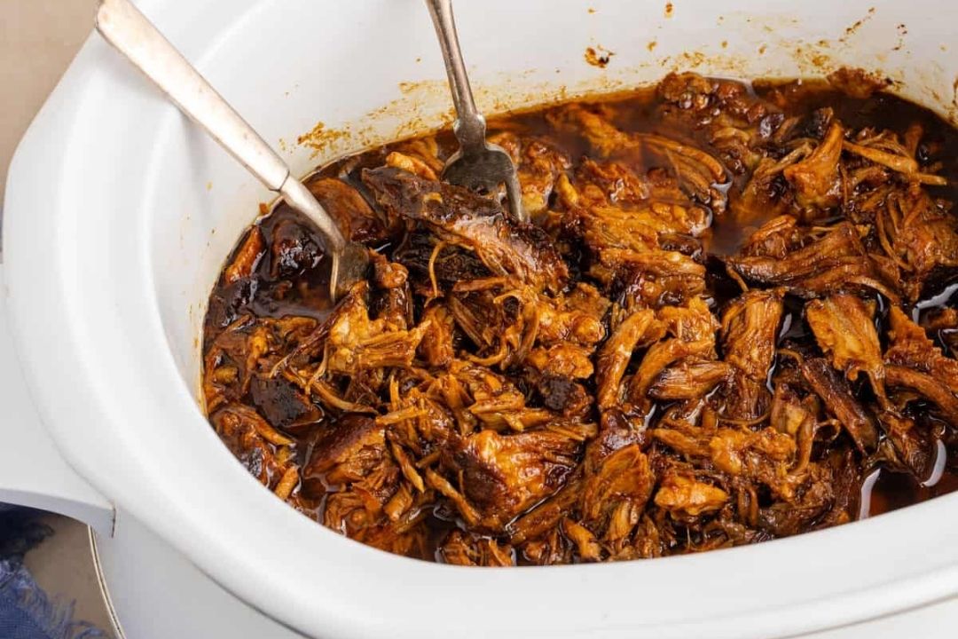 Shredded, saucy beef in a white slow cooker with two metal serving utensils resting inside—an inviting dish perfect for cozy fall recipes and crowd-pleasing Slow Cooker gatherings.