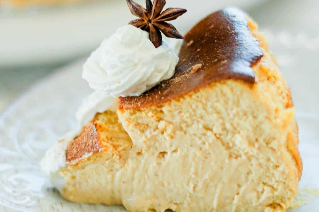 A slice of creamy cheesecake with a browned top, inspired by the cozy flavors of pumpkin season, topped with a swirl of whipped cream and a star anise, served on a white plate.