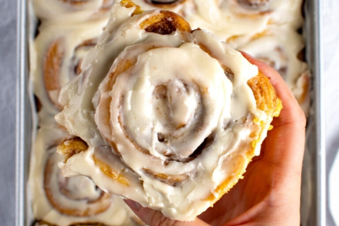 A hand holds a frosted cinnamon roll above a tray filled with more cinnamon rolls covered in icing, making it the perfect treat to enjoy during pumpkin season and savor alongside your favorite fall recipes.