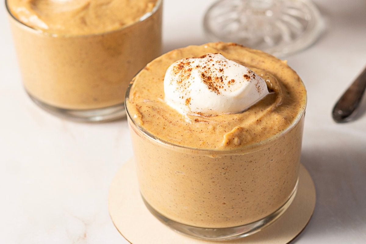 Two glass cups filled with pumpkin mousse, one topped with a dollop of whipped cream and sprinkled with cinnamon, make a delightful addition to your collection of fall recipes on a white surface.