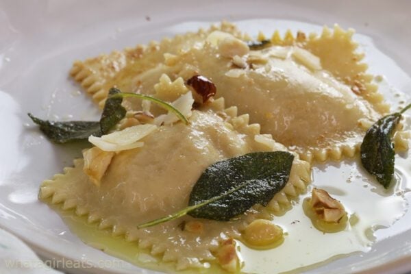 Two ravioli pieces, perfect for pumpkin season, are topped with toasted nuts, cheese shavings, and fried sage leaves, served in a light butter sauce on a white plate.