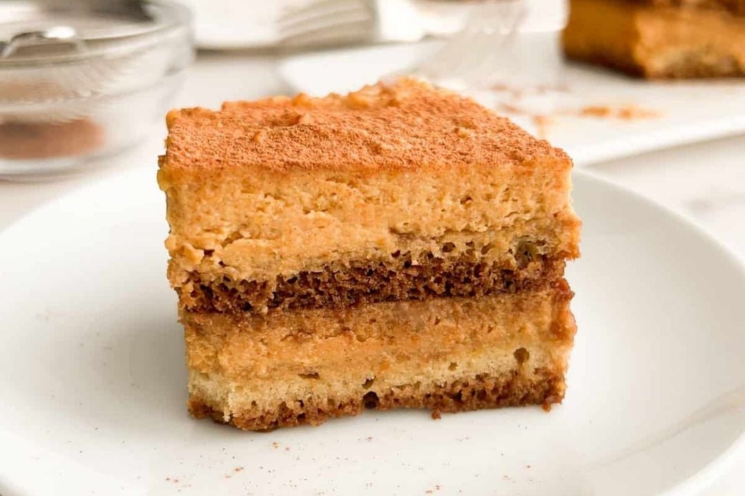 A square slice of layered tiramisu with a dusting of cocoa powder on top is served on a white plate, making it a delightful twist for pumpkin season.