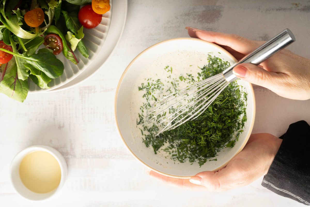 A person whisking chopped herbs into a creamy mixture in a bowl, with a bowl of salad greens and a small bowl of homemade dressing nearby on a white surface.
